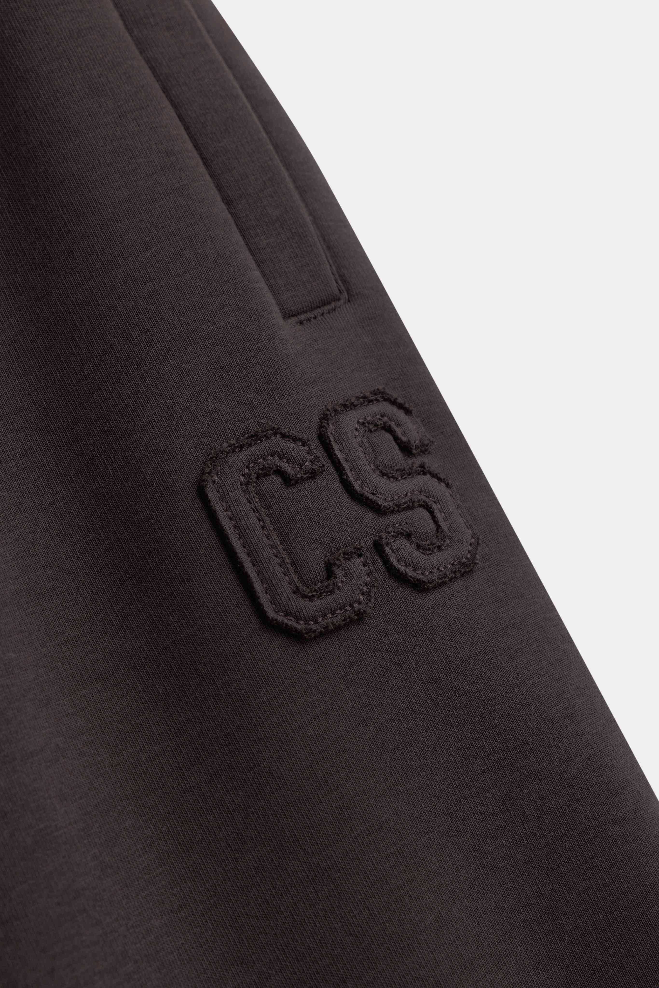 Close-up of espresso CASPARA sweatpants showing the tonal CS logo embroidery and pocket detail.